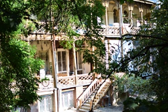 A wooden staircase leads up to a veranda with intricate railing designs, surrounded by lush green foliage. The building features large windows and a second-story balcony adorned with ornate columns and a sunlit seating area.