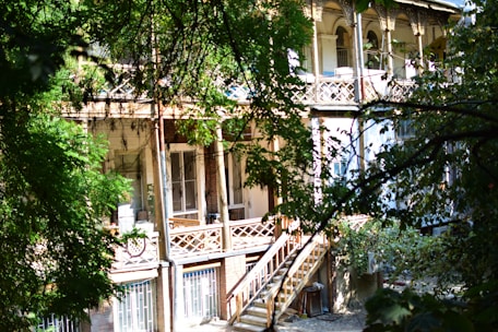 A wooden staircase leads up to a veranda with intricate railing designs, surrounded by lush green foliage. The building features large windows and a second-story balcony adorned with ornate columns and a sunlit seating area.