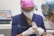 A dental professional wearing a pink cap and protective eyewear is performing a procedure using a specialized instrument. They are in a clinical setting, with a computer displaying dental imagery in the background. Another person, likely a patient, wearing a colorful cap, is partially visible.