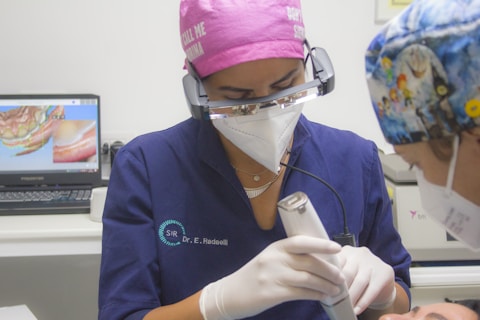 A dental professional wearing a pink cap and protective eyewear is performing a procedure using a specialized instrument. They are in a clinical setting, with a computer displaying dental imagery in the background. Another person, likely a patient, wearing a colorful cap, is partially visible.