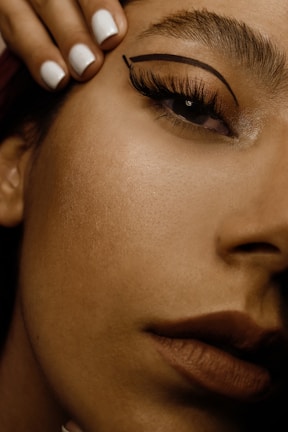 A close-up of a woman's face focusing on the eye area, showcasing detailed makeup with bold eyeliner and well-groomed eyebrows. Her lips are visible in the bottom right, with a natural-looking lipstick. A hand with neatly manicured white nails is positioned near her forehead.