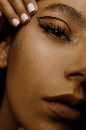 A close-up of a woman's face focusing on the eye area, showcasing detailed makeup with bold eyeliner and well-groomed eyebrows. Her lips are visible in the bottom right, with a natural-looking lipstick. A hand with neatly manicured white nails is positioned near her forehead.