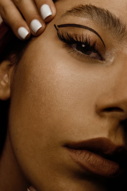 A close-up of a woman's face focusing on the eye area, showcasing detailed makeup with bold eyeliner and well-groomed eyebrows. Her lips are visible in the bottom right, with a natural-looking lipstick. A hand with neatly manicured white nails is positioned near her forehead.