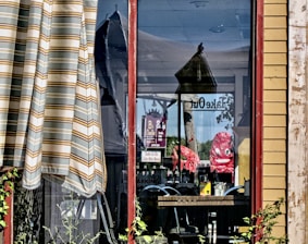 A window reflection reveals various items, including red chairs, tables, and a sign that says 'Take Out.' Outside, there is a striped curtain and some greenery. The interior appears to be a cozy dining setting.