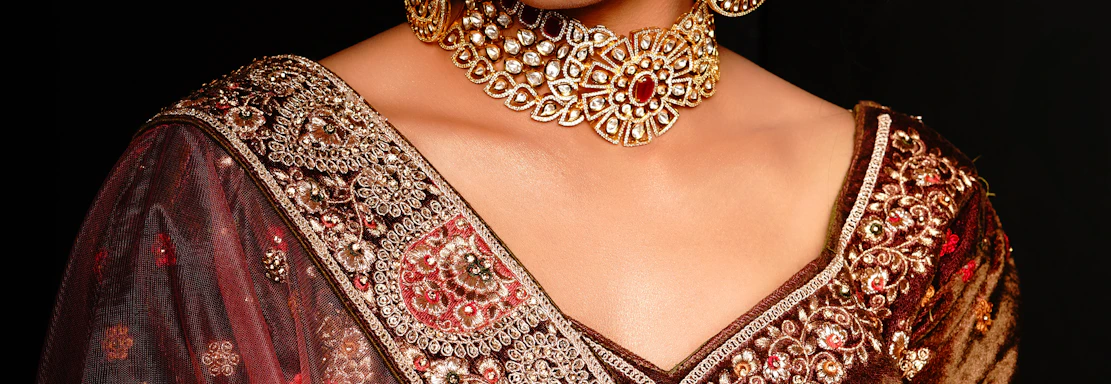 A woman wearing ornate traditional Indian attire with intricate embroidery and embellishments. She has a bejeweled necklace, earrings, and headpiece that match the detailed designs on her clothing. Her expression is calm and composed, with arms crossed gently. The background is dark, enhancing the rich colors and textures of her outfit.