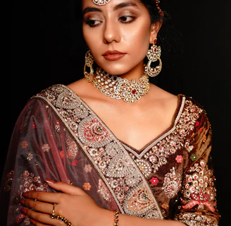 A woman wearing ornate traditional Indian attire with intricate embroidery and embellishments. She has a bejeweled necklace, earrings, and headpiece that match the detailed designs on her clothing. Her expression is calm and composed, with arms crossed gently. The background is dark, enhancing the rich colors and textures of her outfit.