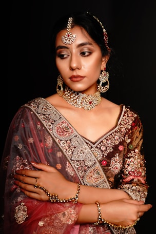 A woman wearing ornate traditional Indian attire with intricate embroidery and embellishments. She has a bejeweled necklace, earrings, and headpiece that match the detailed designs on her clothing. Her expression is calm and composed, with arms crossed gently. The background is dark, enhancing the rich colors and textures of her outfit.