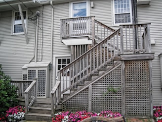 A residential exterior with light gray wooden siding and multiple white-framed windows. A wooden staircase with a weathered appearance leads to an upper door, featuring lattice work beneath and on the side. Colorful flower beds with pink, white, and red blooms are visible near the base of the stairs, adding a vibrant contrast to the muted tones of the building.