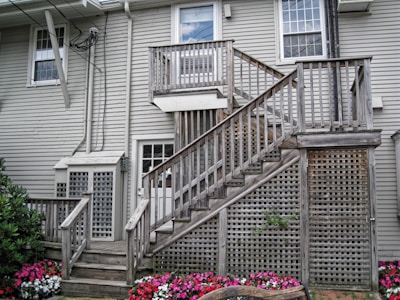A residential exterior with light gray wooden siding and multiple white-framed windows. A wooden staircase with a weathered appearance leads to an upper door, featuring lattice work beneath and on the side. Colorful flower beds with pink, white, and red blooms are visible near the base of the stairs, adding a vibrant contrast to the muted tones of the building.