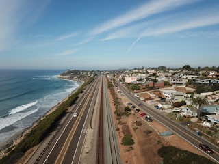 An aerial view of a coastal highway running parallel to the ocean on the left, with waves gently hitting the shore. To the right, there is a residential area with numerous houses and streets. The landscape features a blend of natural coastal beauty and urban development under a clear blue sky.