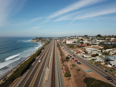 An aerial view of a coastal highway running parallel to the ocean on the left, with waves gently hitting the shore. To the right, there is a residential area with numerous houses and streets. The landscape features a blend of natural coastal beauty and urban development under a clear blue sky.