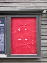 A framed section on a textured wooden wall displaying a panel with a red fabric backdrop. The fabric is slightly wrinkled at various points, and there are scraps of white paper or tape adhered in a random pattern. To the left, a smaller section displays columns of text on a dark blue background.