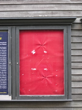 A framed section on a textured wooden wall displaying a panel with a red fabric backdrop. The fabric is slightly wrinkled at various points, and there are scraps of white paper or tape adhered in a random pattern. To the left, a smaller section displays columns of text on a dark blue background.