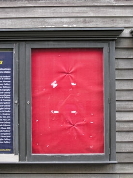 A framed section on a textured wooden wall displaying a panel with a red fabric backdrop. The fabric is slightly wrinkled at various points, and there are scraps of white paper or tape adhered in a random pattern. To the left, a smaller section displays columns of text on a dark blue background.