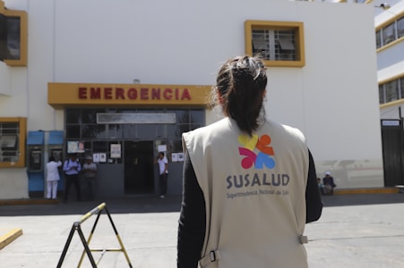 A person wearing a vest with the logo of SUSALUD is standing in front of a hospital emergency entrance. The building is large and painted in white and yellow with a sign labeled 'EMERGENCIA' above the door. There are a few people near the entrance, including what appears to be medical staff.