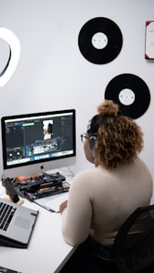 A person with curly hair wearing headphones is sitting at a desk, focused on editing video content on a computer. The desk is equipped with various gadgets, including a laptop and other tech accessories. The room's wall displays vinyl records, adding an artistic touch.