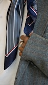 A close-up view of a person's hand with rings, holding the lapel of a grey tweed coat. Two silk ties with patterns, one with stripes and another with paisley designs, are visible. The background is a plain white fabric.