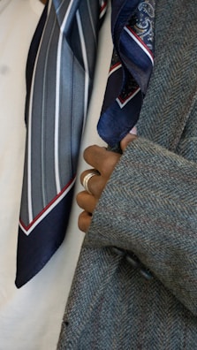 A close-up view of a person's hand with rings, holding the lapel of a grey tweed coat. Two silk ties with patterns, one with stripes and another with paisley designs, are visible. The background is a plain white fabric.
