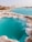 Turquoise water pools bordered by white and brown salt formations create a striking contrast against the barren landscape. The uneven terrain surrounds the vivid blue water, with mounds of earth visible in the background under a clear sky.
