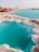Turquoise water pools bordered by white and brown salt formations create a striking contrast against the barren landscape. The uneven terrain surrounds the vivid blue water, with mounds of earth visible in the background under a clear sky.
