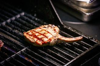 A grilled tomahawk steak with visible char marks sits on a black metal grill. The meat appears juicy and perfectly cooked, with a bone protruding from one end. The grill bars are dark, with hints of red from the fire beneath.