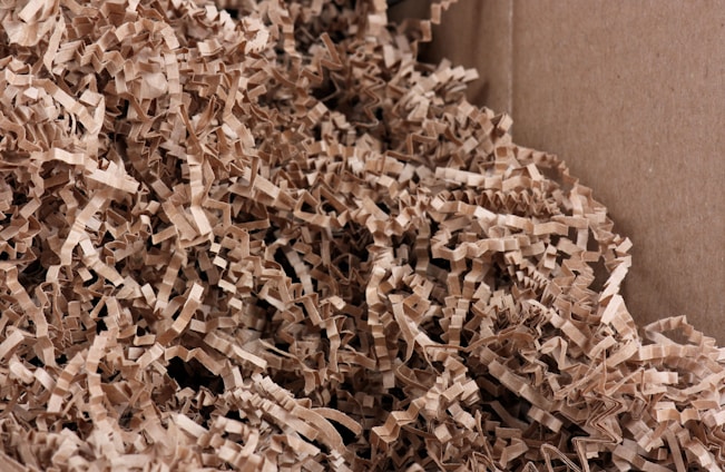 A close-up view of crinkled brown paper strips filling a cardboard box. The paper strips are densely packed and create a textured and layered appearance.