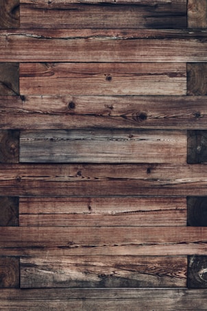 An arrangement of wooden planks with a rustic and weathered appearance. The wood grains and knots are prominently visible, giving it a natural and textured look. The planks are aligned horizontally and vary slightly in color and tone.