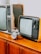 A vintage setting featuring two retro televisions, one larger and one smaller, placed beside a classic analog alarm clock. The items rest on a wooden cabinet, evoking a sense of nostalgia. The televisions have bulky frames and dials typical of older models.