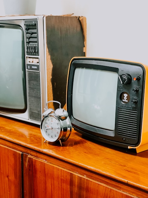 A vintage setting featuring two retro televisions, one larger and one smaller, placed beside a classic analog alarm clock. The items rest on a wooden cabinet, evoking a sense of nostalgia. The televisions have bulky frames and dials typical of older models.