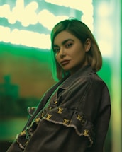 A person with short hair poses confidently in a stylish jacket adorned with designer logos. The background features a vibrant green neon light, creating a modern and trendy atmosphere.