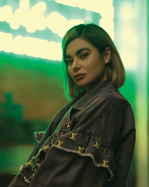 A person with short hair poses confidently in a stylish jacket adorned with designer logos. The background features a vibrant green neon light, creating a modern and trendy atmosphere.