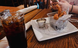 A wooden table at a restaurant is set with a glass of soda, salt and pepper shakers, a basket containing fried food on wax paper styled like newspaper, and a small container of dipping sauce. A person is using a smartphone in the background.