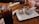 A wooden table at a restaurant is set with a glass of soda, salt and pepper shakers, a basket containing fried food on wax paper styled like newspaper, and a small container of dipping sauce. A person is using a smartphone in the background.