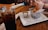 A wooden table at a restaurant is set with a glass of soda, salt and pepper shakers, a basket containing fried food on wax paper styled like newspaper, and a small container of dipping sauce. A person is using a smartphone in the background.