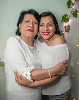 Two women are standing close together, smiling at the camera. They are embracing each other warmly. Both women are wearing white tops. There are fairy lights and green leaves in the background.