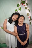 Two young girls are standing close together, embracing each other with smiles. They are dressed in formal attire; one in a white dress and the other in a dark blue dress. Behind them is a decorative background with green leaves, red flowers, and string lights forming an arch shape.