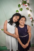 Two young girls are standing close together, embracing each other with smiles. They are dressed in formal attire; one in a white dress and the other in a dark blue dress. Behind them is a decorative background with green leaves, red flowers, and string lights forming an arch shape.