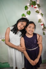 Two young girls are standing close together, embracing each other with smiles. They are dressed in formal attire; one in a white dress and the other in a dark blue dress. Behind them is a decorative background with green leaves, red flowers, and string lights forming an arch shape.