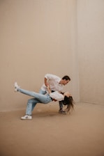A couple is engaged in a dance pose, with the man supporting the woman as she leans back, her hair flowing freely. The setting is minimal, with light-colored walls and flooring.
