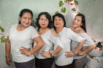 Four women are standing in a line, each posing with one hand on their hip. They are all smiling and wearing white tops. The background features twinkling lights and some greenery.