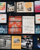 A collection of various photography books displayed on two black shelves. The covers feature different styles and themes: a Polaroid guide, street photography manuals, and books on the history of photography. Some notable titles include 'The Station' by Chris Killip, 'The Way It Was' by Thomas Hoepker, and a book titled 'Heroes'. Each book cover has distinct graphic designs, typography, and color schemes.