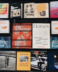 A collection of various photography books displayed on two black shelves. The covers feature different styles and themes: a Polaroid guide, street photography manuals, and books on the history of photography. Some notable titles include 'The Station' by Chris Killip, 'The Way It Was' by Thomas Hoepker, and a book titled 'Heroes'. Each book cover has distinct graphic designs, typography, and color schemes.