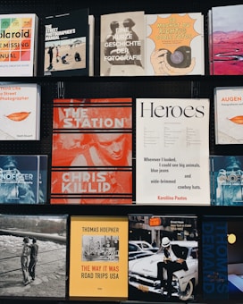 A collection of various photography books displayed on two black shelves. The covers feature different styles and themes: a Polaroid guide, street photography manuals, and books on the history of photography. Some notable titles include 'The Station' by Chris Killip, 'The Way It Was' by Thomas Hoepker, and a book titled 'Heroes'. Each book cover has distinct graphic designs, typography, and color schemes.