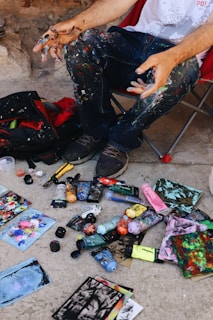 A person sits on a chair with paint-splattered clothing, surrounded by an array of paint tubes, brushes, and colorful paintings scattered on the ground. The person's hands are covered in paint, indicating active involvement in creating artwork.