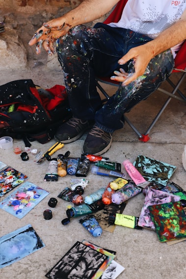 A person sits on a chair with paint-splattered clothing, surrounded by an array of paint tubes, brushes, and colorful paintings scattered on the ground. The person's hands are covered in paint, indicating active involvement in creating artwork.