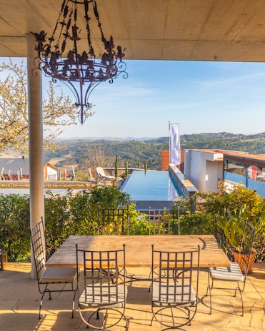 A scenic outdoor setting featuring a wooden table with six wrought iron chairs on a patio. Above, a decorative chandelier hangs from the ceiling. In the background, an infinity pool stretches towards a scenic view of rolling hills and a clear blue sky. Lush greenery surrounds the patio area, enhancing the tranquil atmosphere.