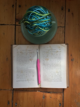 A wooden surface holds an open book with a pink-handled crochet hook placed horizontally across its pages. Nearby, a green plate supports a ball of blue and green yarn.