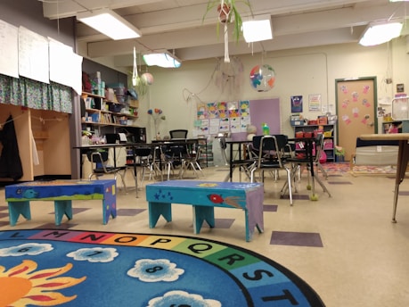 A colorful classroom with small tables and chairs, bright decorations, and educational materials. Children's artwork is displayed on the walls, and there are playful elements like painted benches and a vibrant alphabet rug.