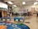 A colorful classroom with small tables and chairs, bright decorations, and educational materials. Children's artwork is displayed on the walls, and there are playful elements like painted benches and a vibrant alphabet rug.