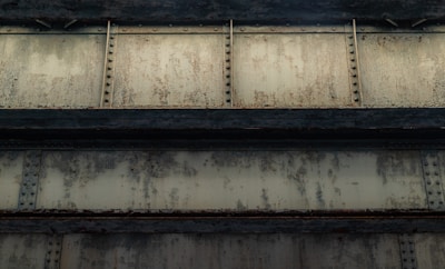 A close-up view of an industrial metal structure featuring rusted steel beams with rivets and a weathered, textured surface. The beams have a greenish, corroded appearance, suggesting wear and age.
